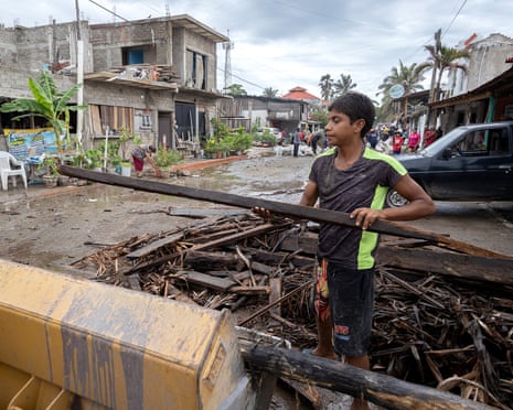 Boy in green and black T-shirt lifting a beam of wood from wreckage on a muddy street.